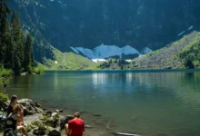 A panoramic view of snow-capped mountains reflecting on a serene alpine lake under a clear blue sky.