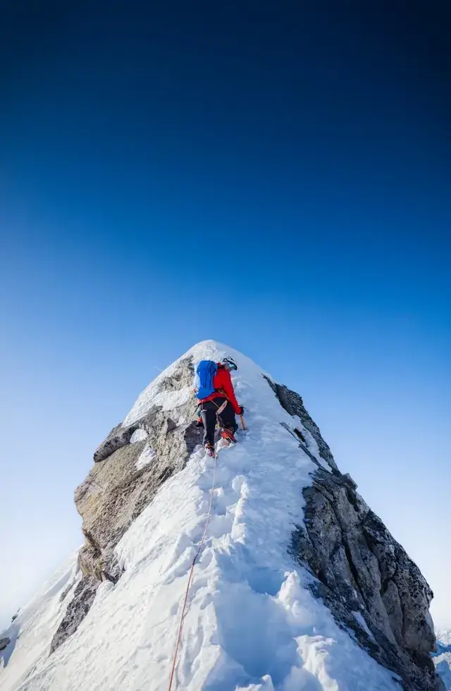 A mountaineer triumphantly stands atop a snow-covered peak, raising a flag against a backdrop of clear blue skies and rugged mountain ranges.